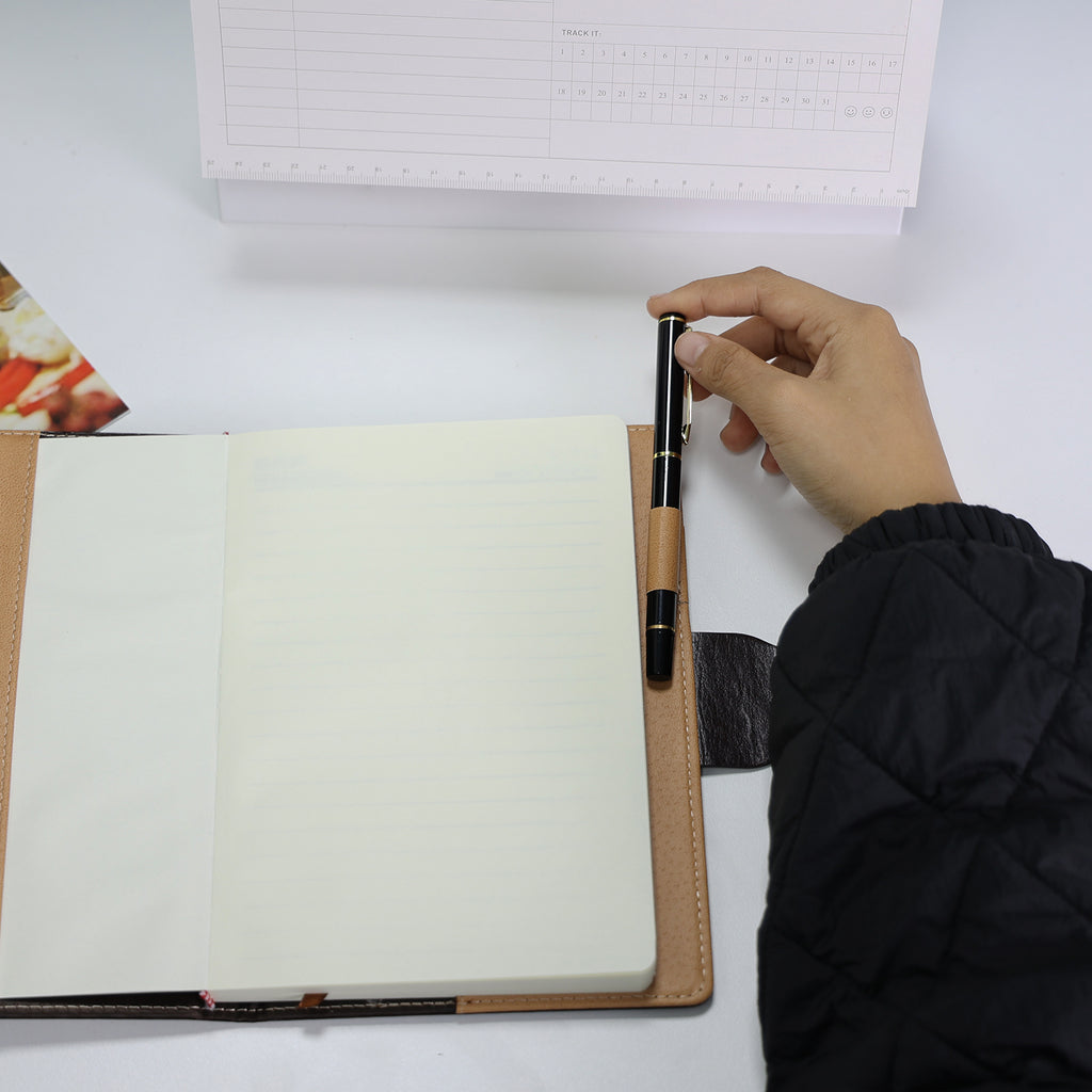 Foto do Caderno com Cadeado Cartagena LUMAI preto aberto, com mão segurando caneta sobre páginas em branco, mostrando as páginas internas.