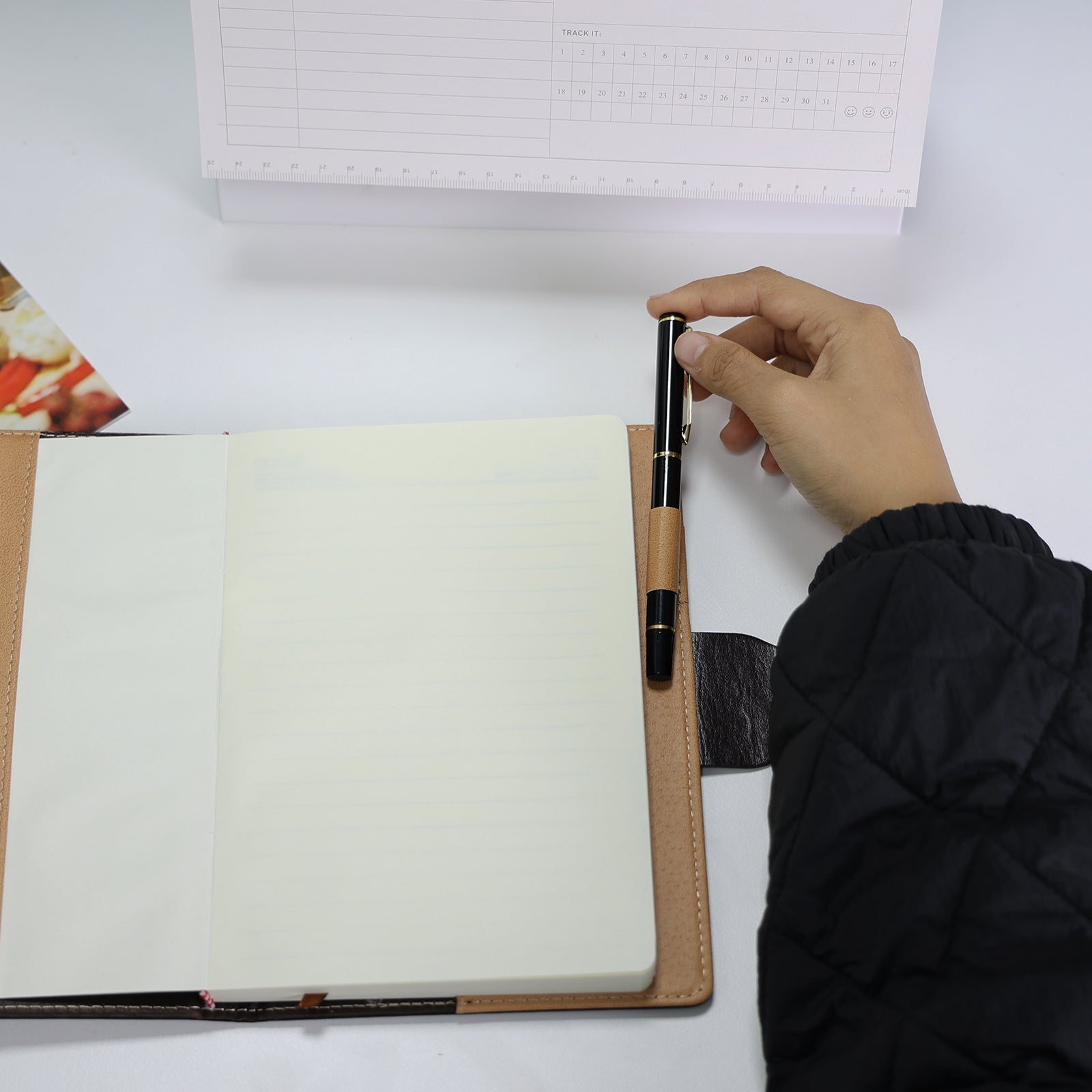 Foto do Caderno com Cadeado Cartagena LUMAI preto aberto, com mão segurando caneta sobre páginas em branco, mostrando as páginas internas.