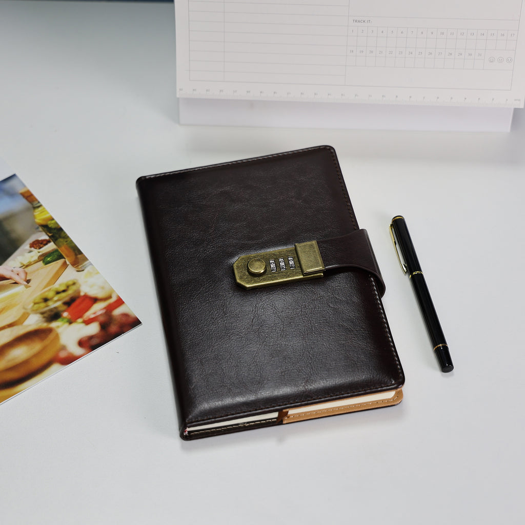 Foto lifestyle do Caderno com Cadeado Cartagena LUMAI preto sobre mesa com caneta e fotos, exibindo a capa de couro sintético com fechadura dourada.