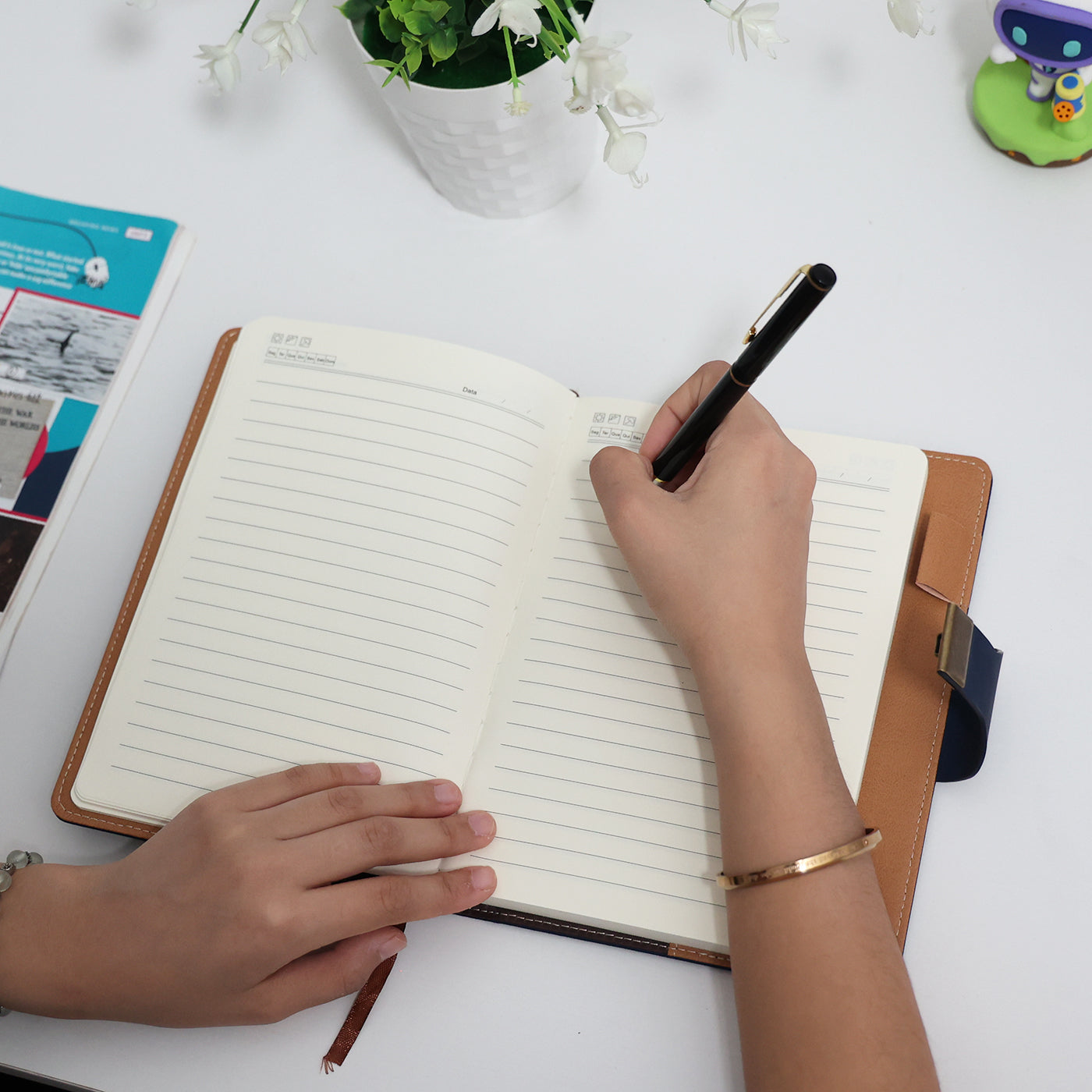 Foto lifestyle de mão feminina escrevendo no caderno diário de couro azul aberto sobre mesa branca com flores e decorações ao fundo, transmitindo sofisticação e uso cotidiano.