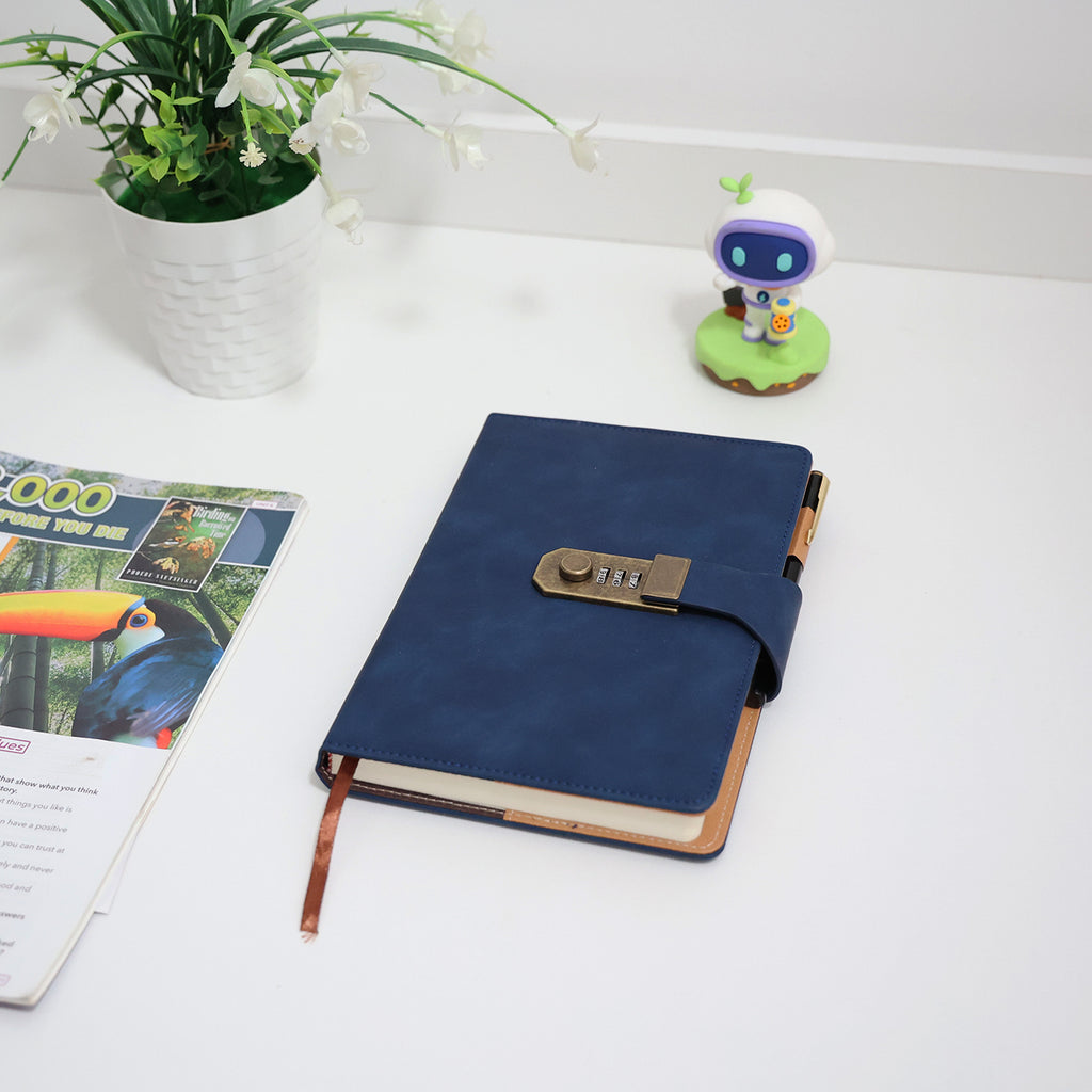 Foto lifestyle do caderno diário de couro azul fechado sobre mesa branca decorada com flores brancas, brinquedo robô e revistas, transmitindo estilo e personalidade.