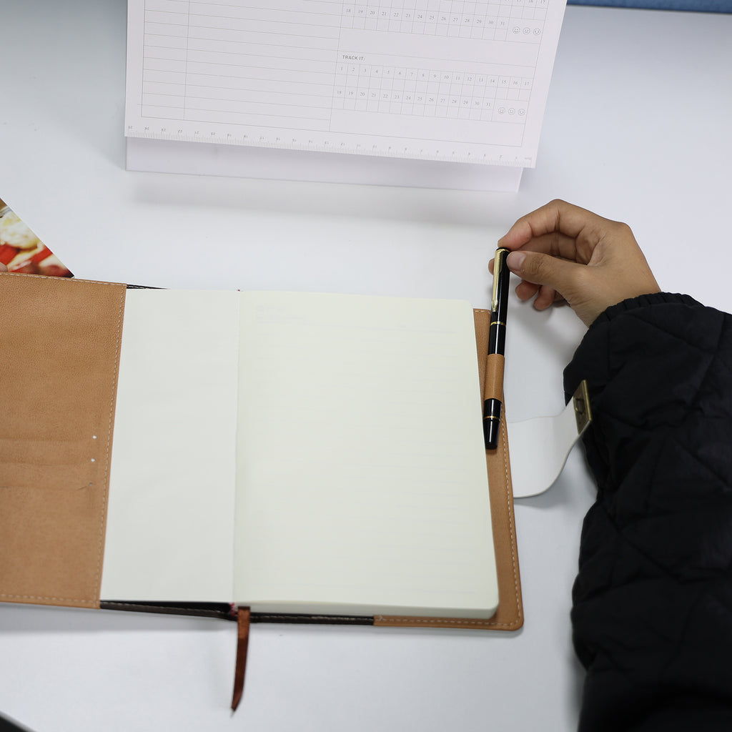 Foto de mão segurando caderno diário de couro branco aberto com páginas em branco e caneta, sobre mesa com fundo floral. Mostra interior e qualidade das páginas do produto.