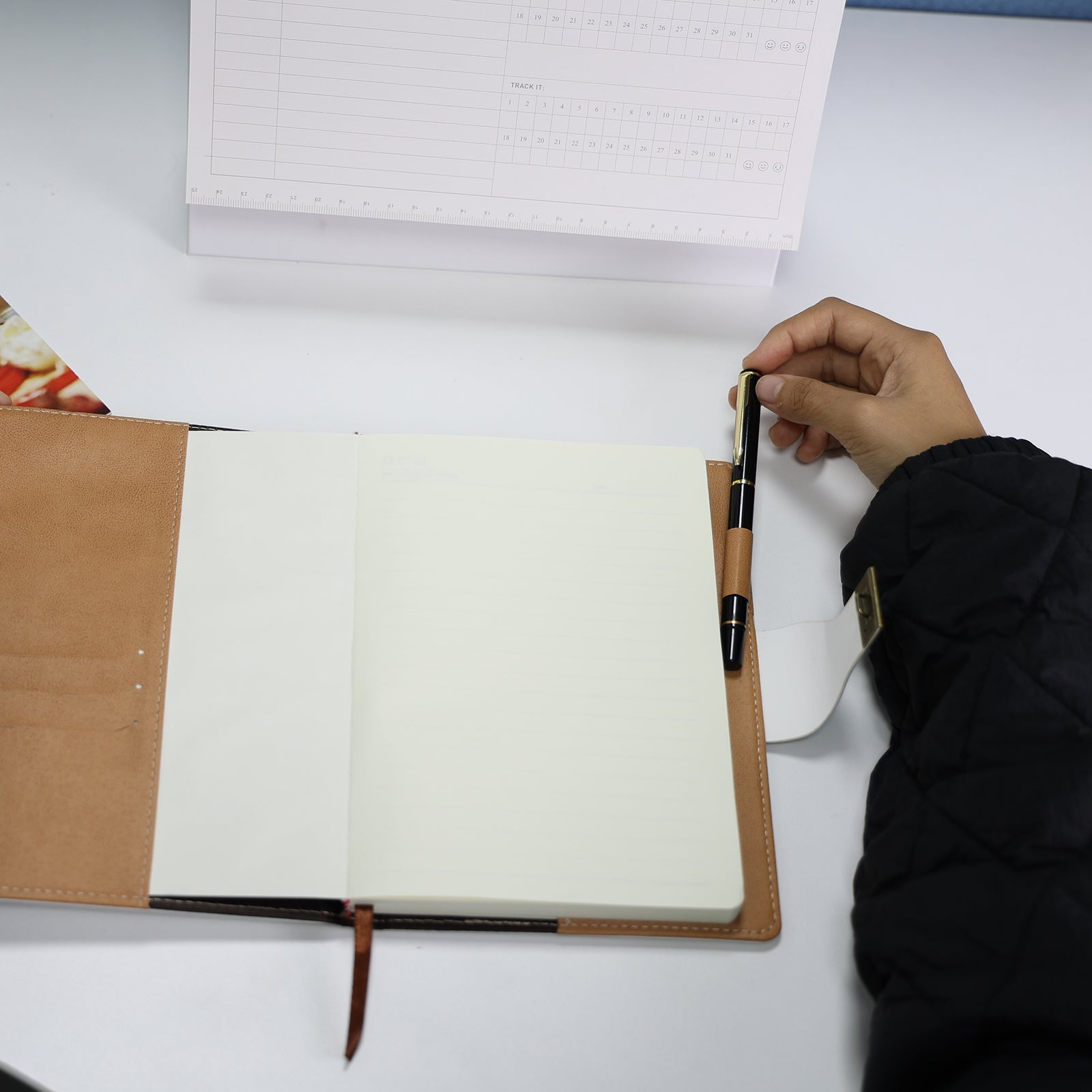 Foto de mão segurando caderno diário de couro branco aberto com páginas em branco e caneta, sobre mesa com fundo floral. Mostra interior e qualidade das páginas do produto.
