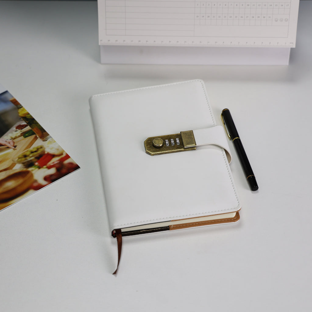 Caderno diário de couro branco com fechamento por cadeado de combinação e caneta preta, fotografado sobre mesa branca com foto decorativa ao fundo.