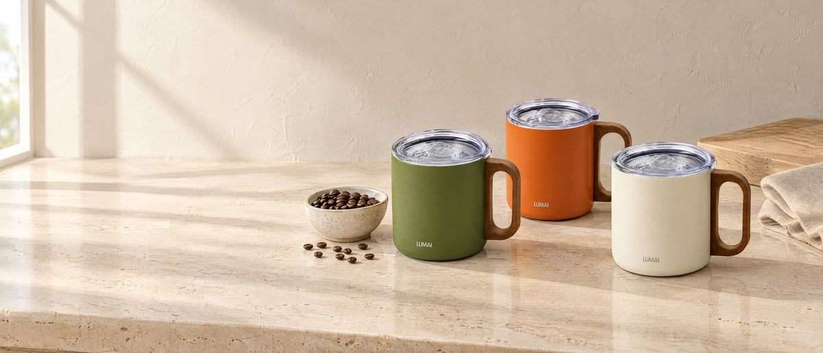 Canecas Caraíva verde, laranja e branca, com tampa, sobre uma bancada de pedra clara com grãos de café